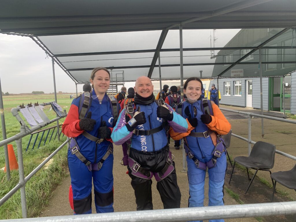 3 people smiling for a group photo wearing skydiving suits ready to participate in a sponsored skydive for charity