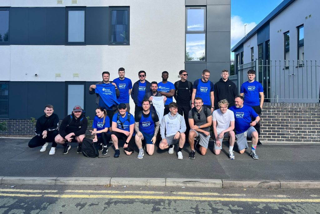 A group photo of a large staff team in charity t-shirts outside of their office building before they walk 32 miles for charity.