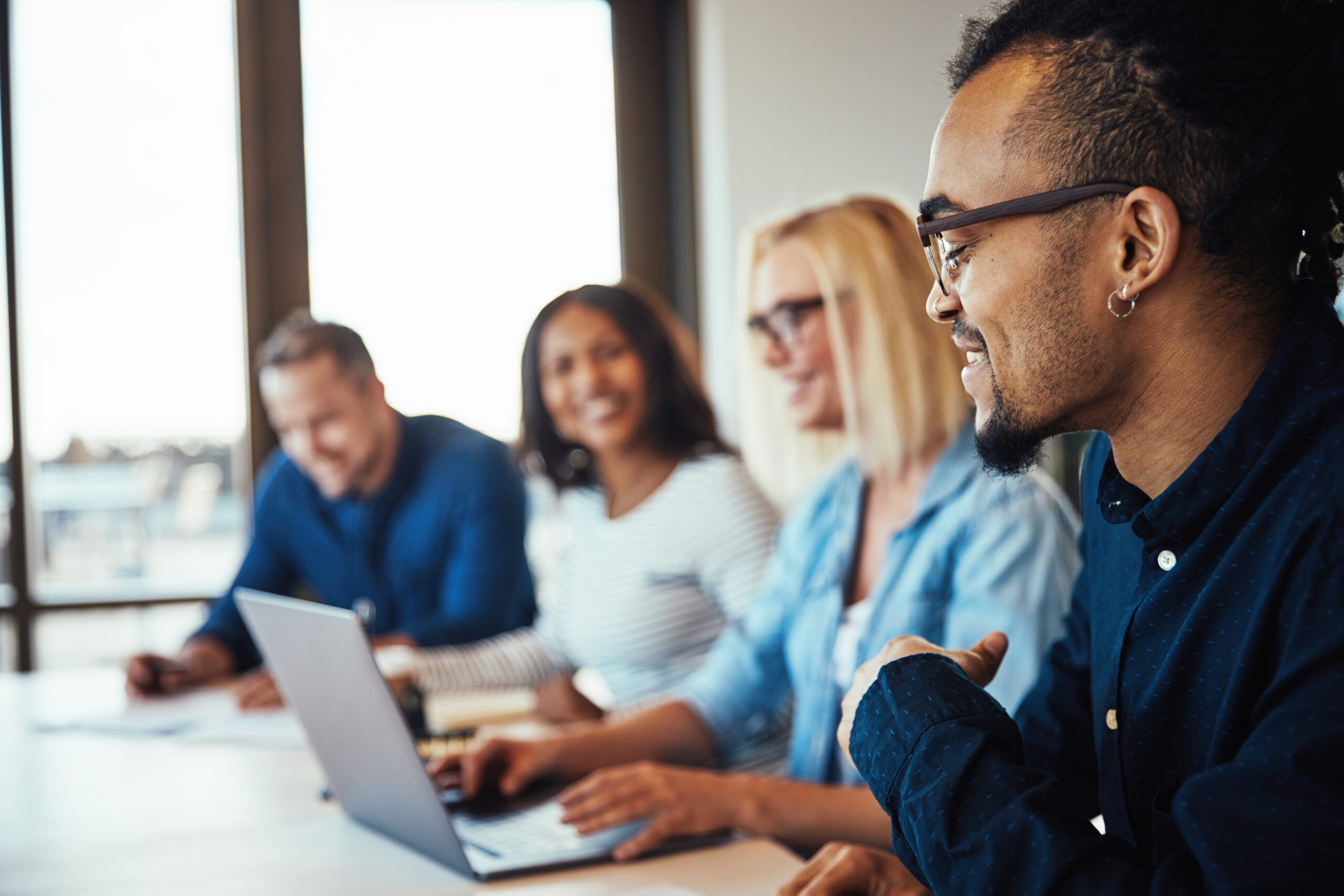 Smiling young African American businessman discussing work while sitting with a diverse group of colleagues during a meeting negotiating salary.